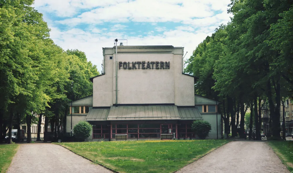 Bild på Folkteatern Gävleborgs huvudentré. På husets står det "FOLKTEATERN" och runtomkring byggnaden finns grönskande träd. Längst ned på bilden breder sig två grusgångar som bildar en triangel som är täckt med gräs.