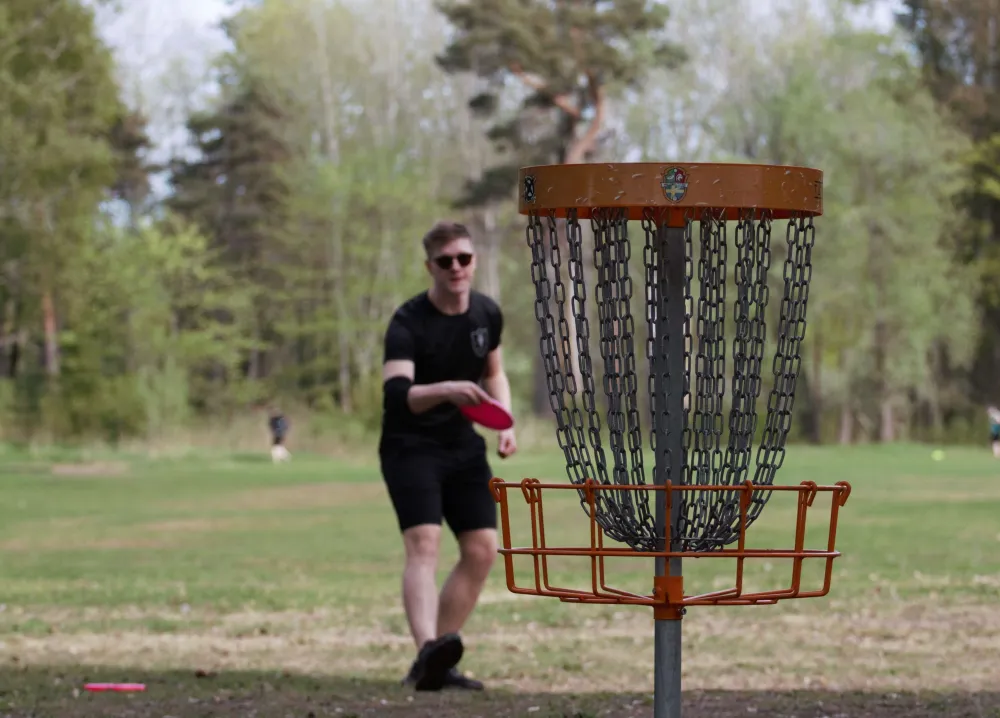 Man som kastar frisbeedisc med en discgolfkorg i förgrunden.